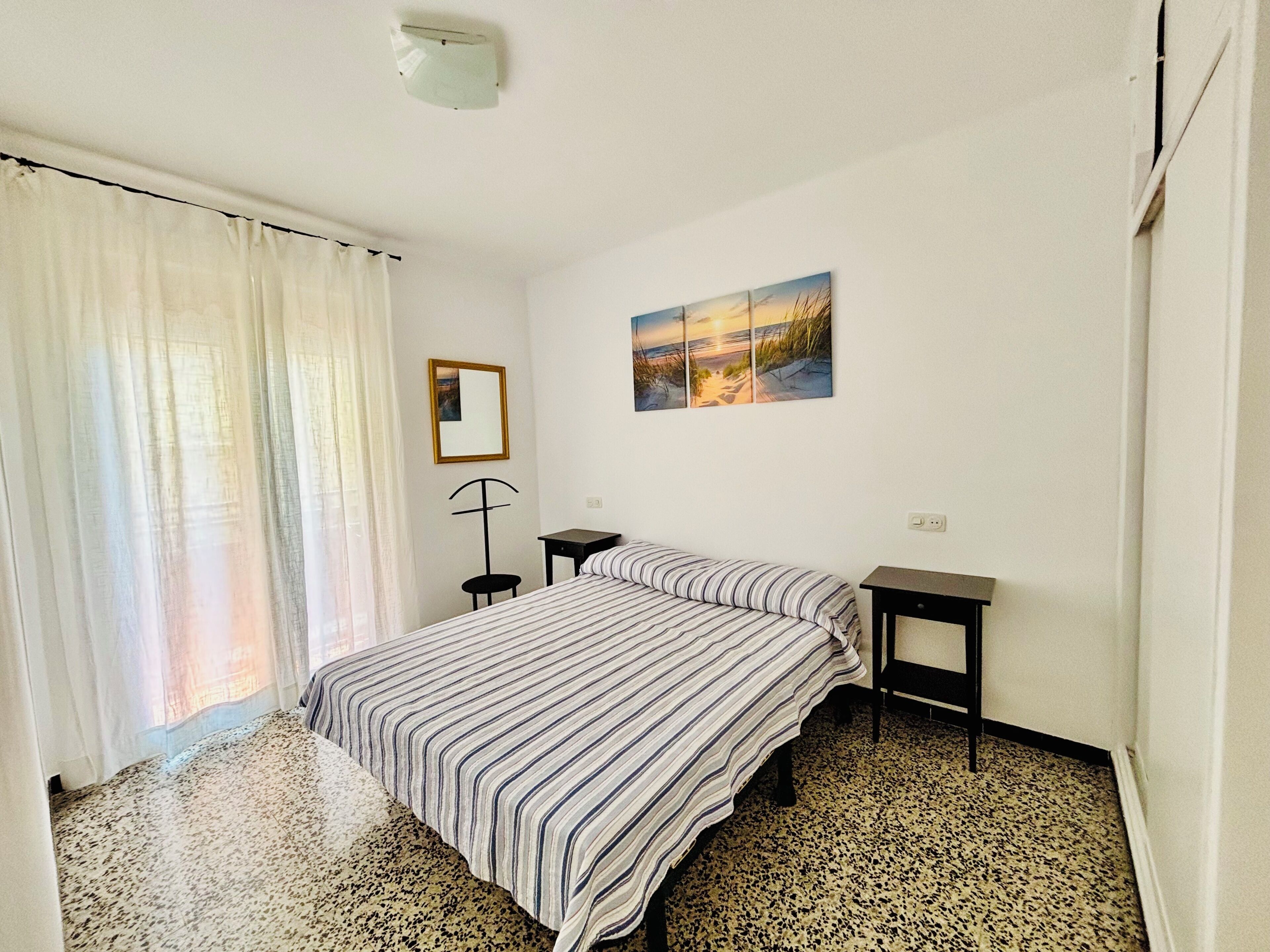 A bedroom with a bed, a nightstand, a floor lamp, a painting, and a window with curtains.
