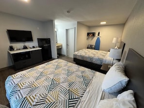Ein Hotelzimmer mit einem großen Bett, einem kleineren Bett, einem Schreibtisch mit Flachbildfernseher, einem Kühlschrank, einem Sessel und einer Lampe.