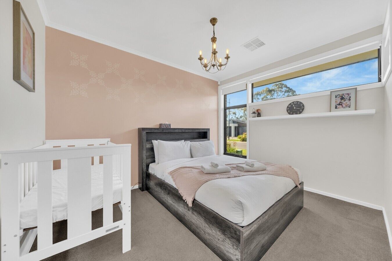 A bedroom with a bed, a crib, a chandelier, a window with a view, and a shelf with clocks.