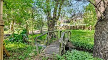 Een houten brug in een bebost gebied, met een boomstam aan de rechterkant en een gebouw op de achtergrond.