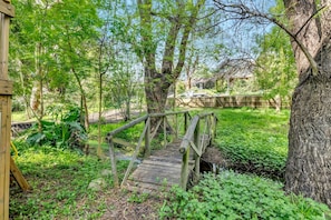 Un pont en bois, situé dans une zone boisée, avec un tronc d’arbre sur la droite et un bâtiment en arrière-plan.