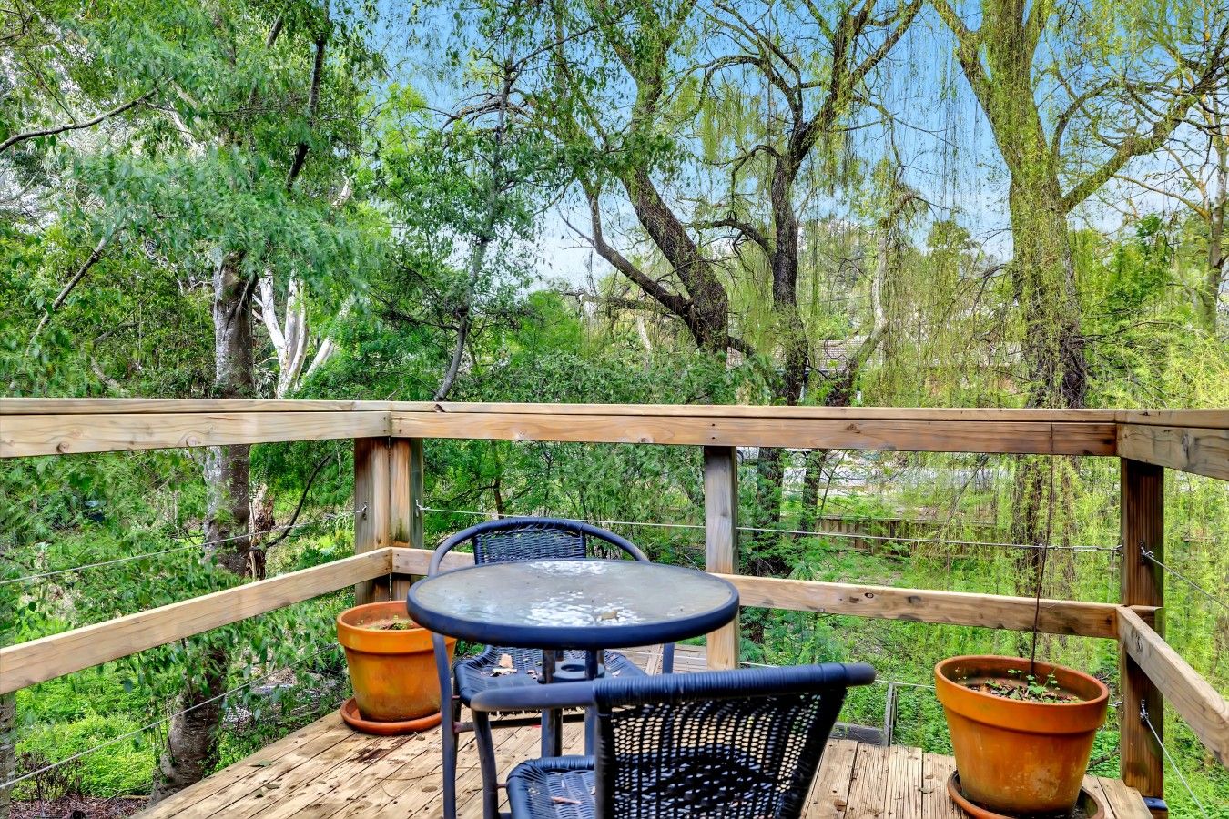 Une terrasse extérieure avec une rambarde en bois, une table ronde, deux chaises et deux plantes en pot.