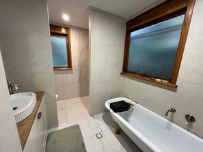 A bathroom with a white bathtub, a wooden vanity, a window, and a tiled floor. - Adelaide Hills Creek-Side Retreat (Bridgewater)