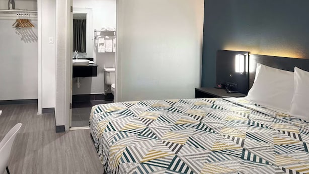 Ein Hotelzimmer mit Bett, Nachttisch, Lampe, Spiegel, Badezimmer mit Waschbecken und Handtuchhalter sowie einer Tür zu einem weiteren Raum.