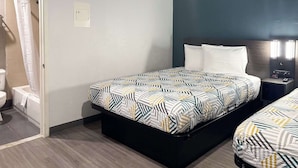 Hotellihuone, jossa on sänky, yöpöytä, seinään kiinnitetty valaisin ja kylpyhuone, joka näkyy avoimesta ovesta.