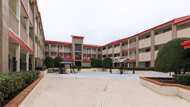 A courtyard with a covered seating area, a small kiosk, and a building with multiple balconies and a red roof. - Enjoy Affordable Comfort | 2 Units in Central Irving | Pet-Friendly Stay (Irving)