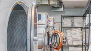 Un cuarto de lavandería con lavadoras y secadoras industriales, estanterías de metal con ropa de cama doblada y piso de baldosas.