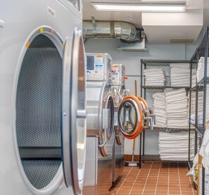 Un cuarto de lavandería con lavadoras y secadoras industriales, estanterías de metal con ropa de cama doblada y piso de baldosas.