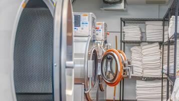 A laundry facility with industrial washers and dryers, metal shelving with folded linens, and a tiled floor.