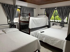 Ein Zimmer mit zwei Einzelbetten und einem Etagenbett, grauen Vorhängen und einem Fenster mit Blick auf Grünflächen.