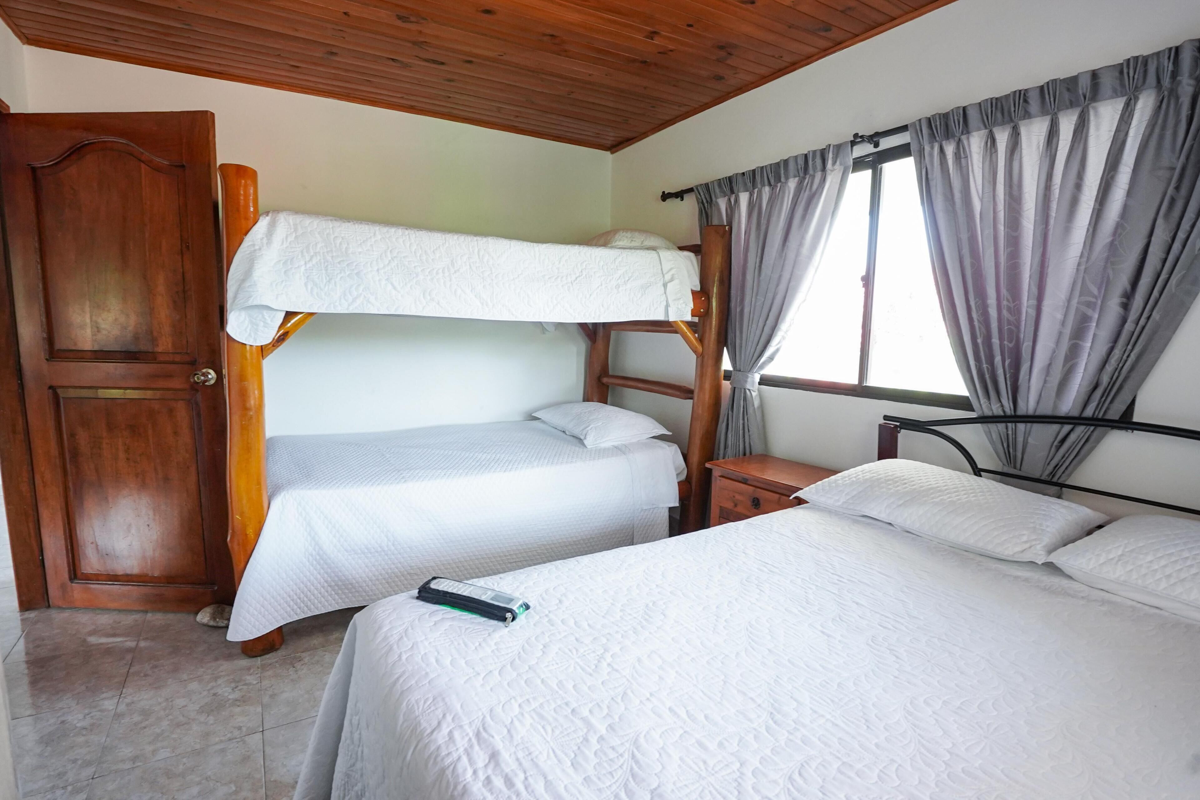 A room with a bunk bed, a single bed, a wooden door, a nightstand, and a window with curtains.