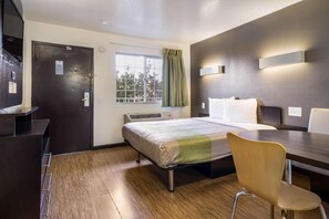 Habitación de hotel con cama, escritorio, silla, ventana con cortinas y lámpara fijada a la pared.