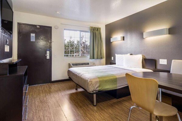 Habitación de hotel con cama, escritorio, silla, ventana con cortinas y lámpara fijada a la pared.