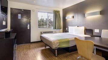 Habitación de hotel con cama, escritorio, silla, ventana con cortinas y lámpara fijada a la pared.