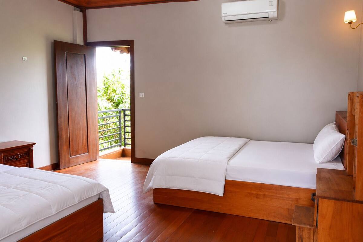 Habitación con dos camas, suelo de madera, armario, aire acondicionado y balcón con plantas.