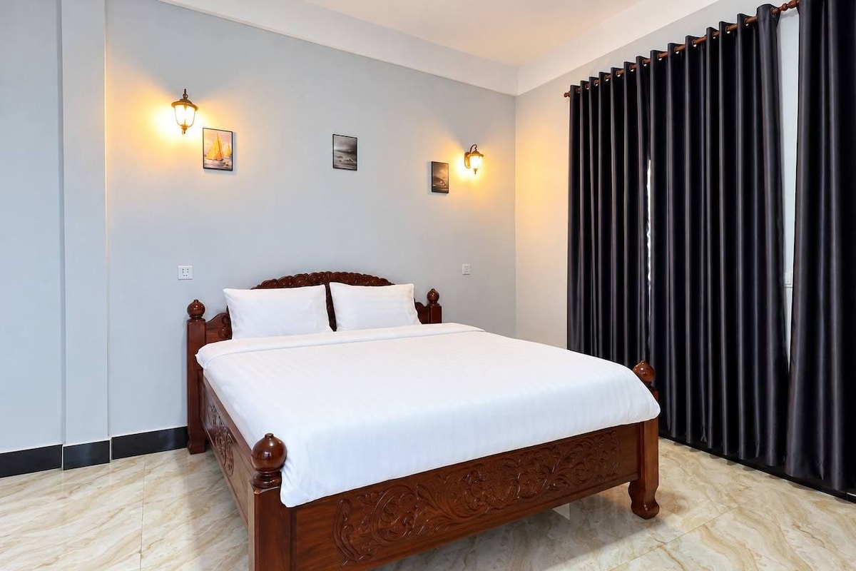 Cama bem arrumada em quarto com piso claro, cortina escura à direita e luminárias fixadas na parede.