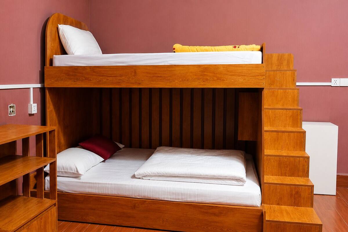 Una cama litera con estructura de madera, colchón blanco y almohada roja. Se observa una repisa de madera con botellas y un pequeño recipiente. También se ve un enchufe de pared blanco.