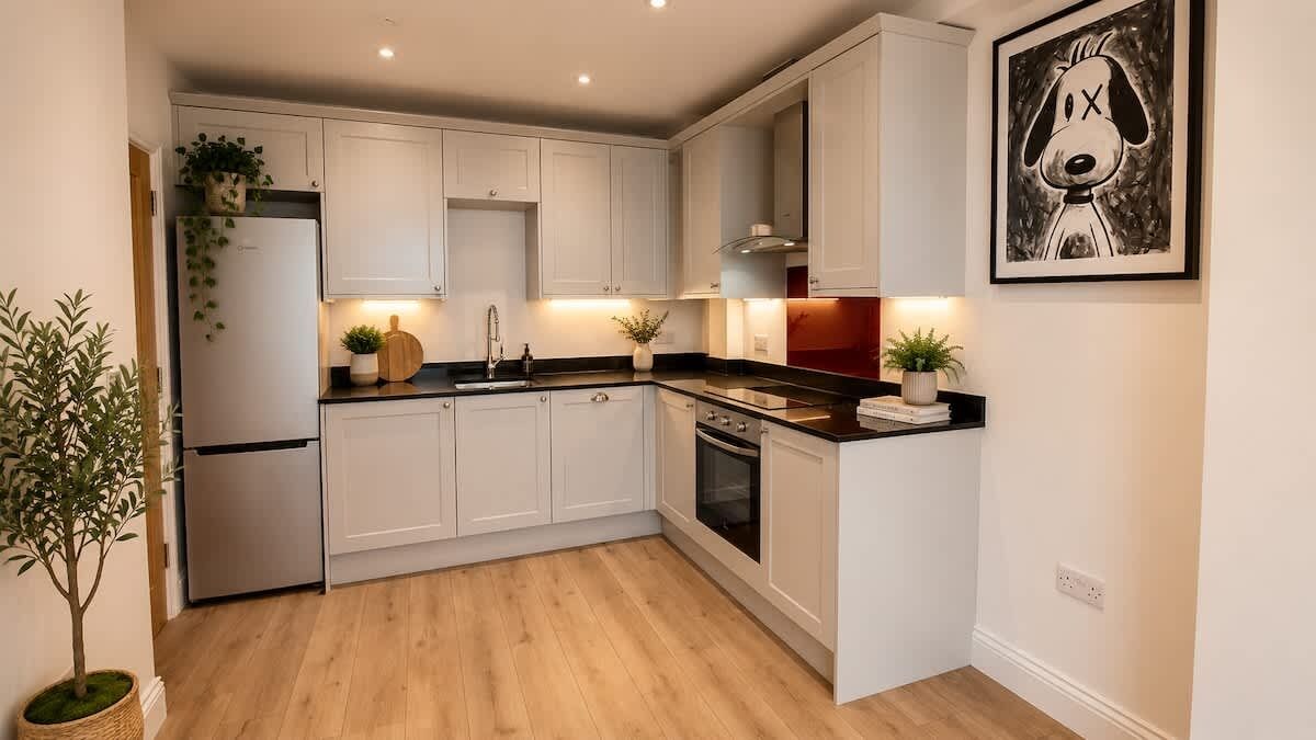 A kitchen with white cabinets, a black countertop, and a refrigerator. There is a plant on the left and a framed picture on the wall.