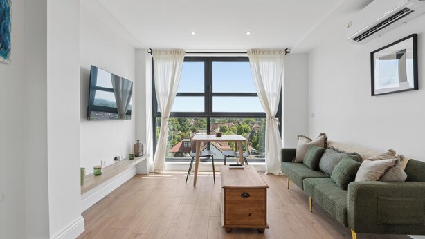 Ein modernes Wohnzimmer mit einem Holztisch, einem grünen Sofa, einem an der Wand montierten Fernseher und einem großen Fenster mit Vorhängen.