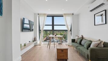 A modern living room with a wooden table, a green sofa, a TV mounted on the wall, and a large window with curtains.