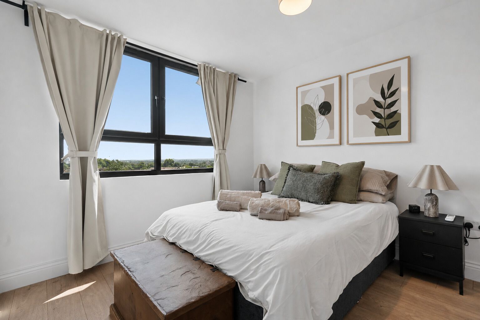 A bedroom with a large bed, two bedside tables, a bench, a window with curtains, and two wall-mounted artworks.