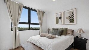 Ein Schlafzimmer mit einem großen Bett, zwei Nachttischen, einer Bank, einem Fenster mit Vorhängen und zwei an der Wand befestigten Kunstwerken.