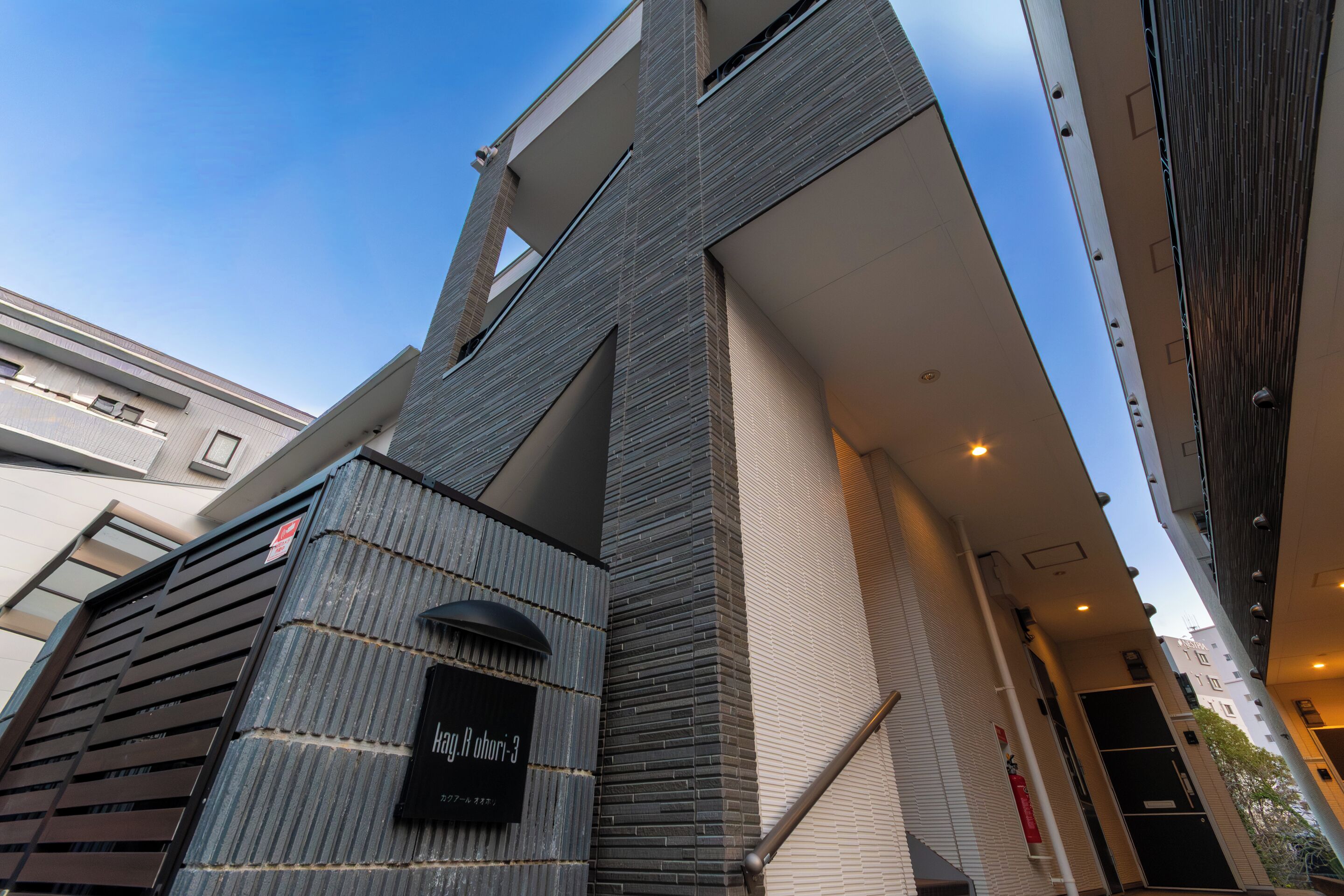 A modern building with a prominent vertical structure, a sign indicating 'Hotel Hohri 3,' and a visible fire extinguisher.