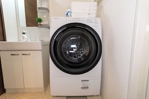 Máquina de lavar com abertura frontal num banheiro compacto e moderno, com pia pequena, espelho e prateleira com produtos de higiene pessoal.