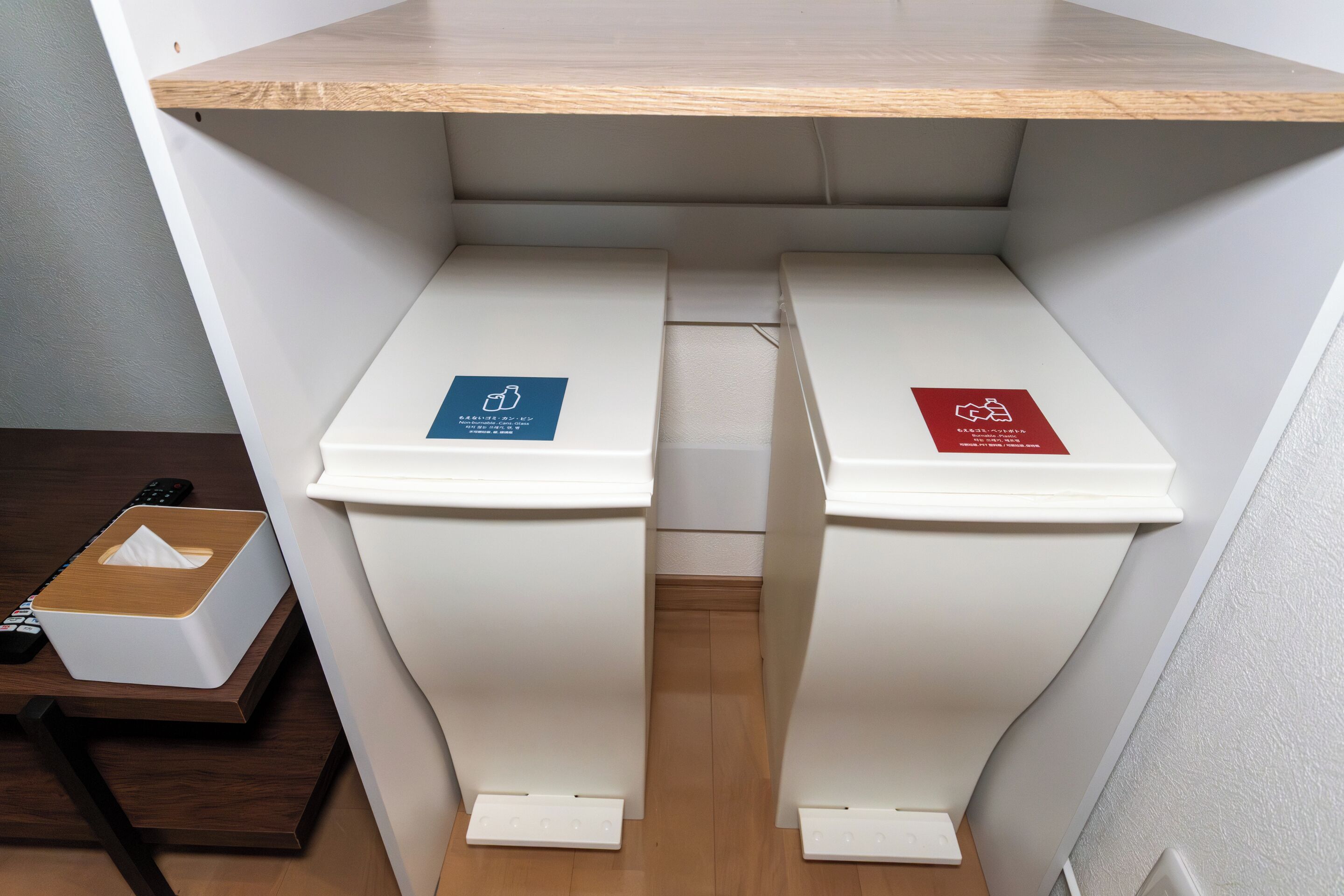 Two white trash bins with blue and red recycling labels, a wooden shelf above, and a remote control on a table.