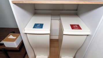 Two white trash bins with blue and red recycling labels, a wooden shelf above, and a remote control on a table.