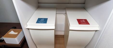 Two white trash bins with blue and red recycling labels, a wooden shelf above, and a remote control on a table.