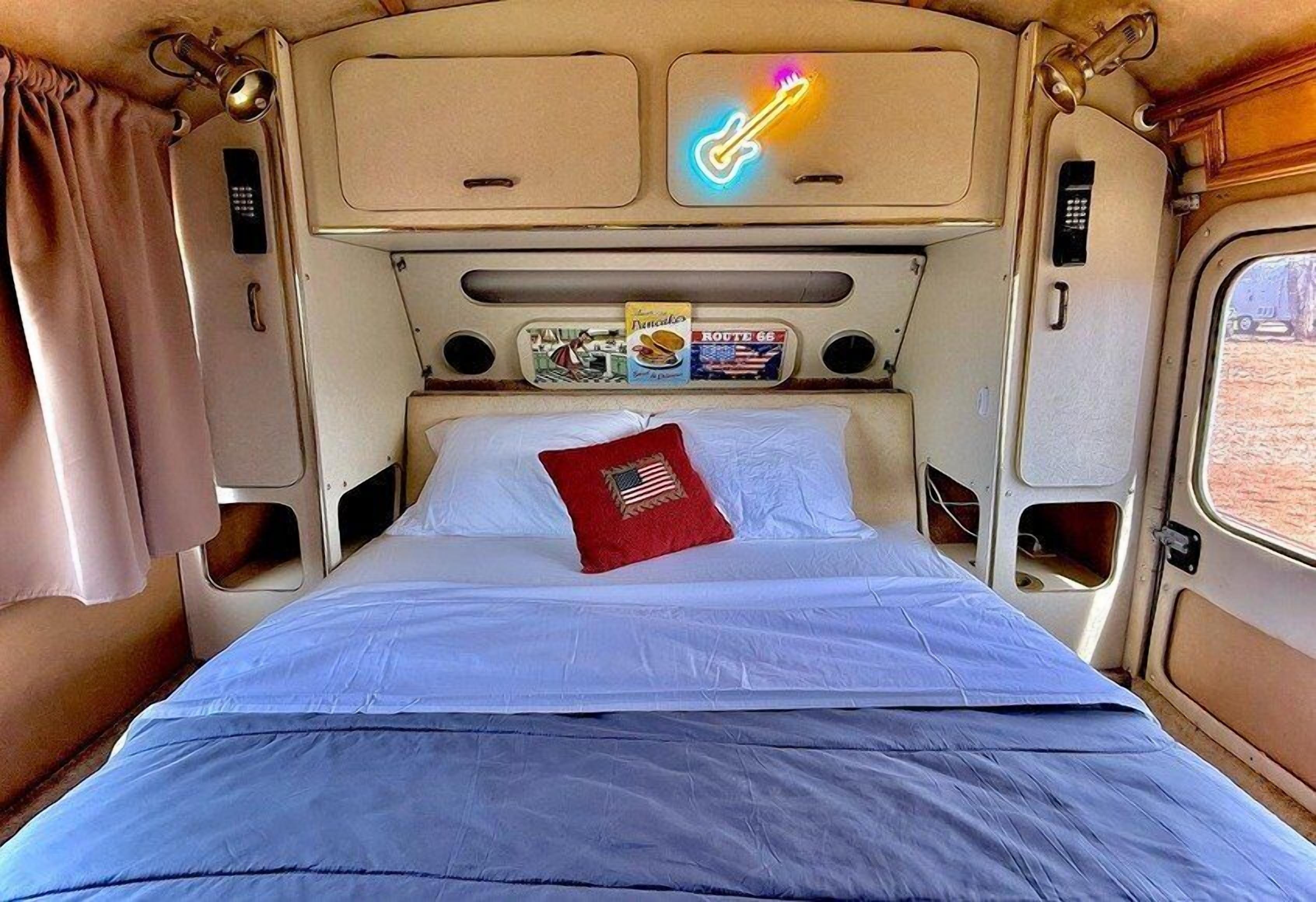 Un llit amb un coixí vermell, un cartell de guitarra de neó i una catifa de revistes a l'interior d'una camper