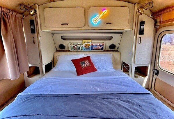 Uma cama com travesseiro vermelho, um letreiro de guitarra neon e um suporte para revistas no interior de uma van camper.