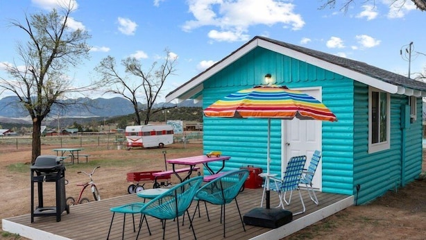 Eine kleine, türkisblaue Holzhütte mit einer Terrasse, Sitzgelegenheiten im Freien und einem farbenfrohen Sonnenschirm.