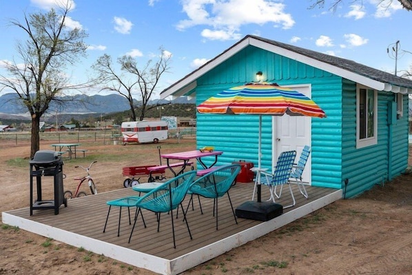 A small turquoise cabin with a deck, outdoor seating, and a colorful umbrella. - Baywatch Cottage | 1 Unit | Near Cañon City Attractions (Canon City)
