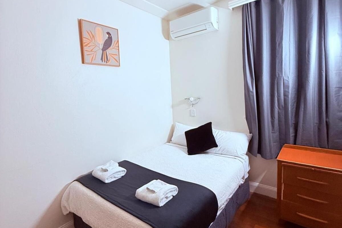 Una habitación de hotel con una cama, una mesita de noche, un equipo de aire acondicionado, una ventana con cortinas y una obra de arte colgada en la pared.