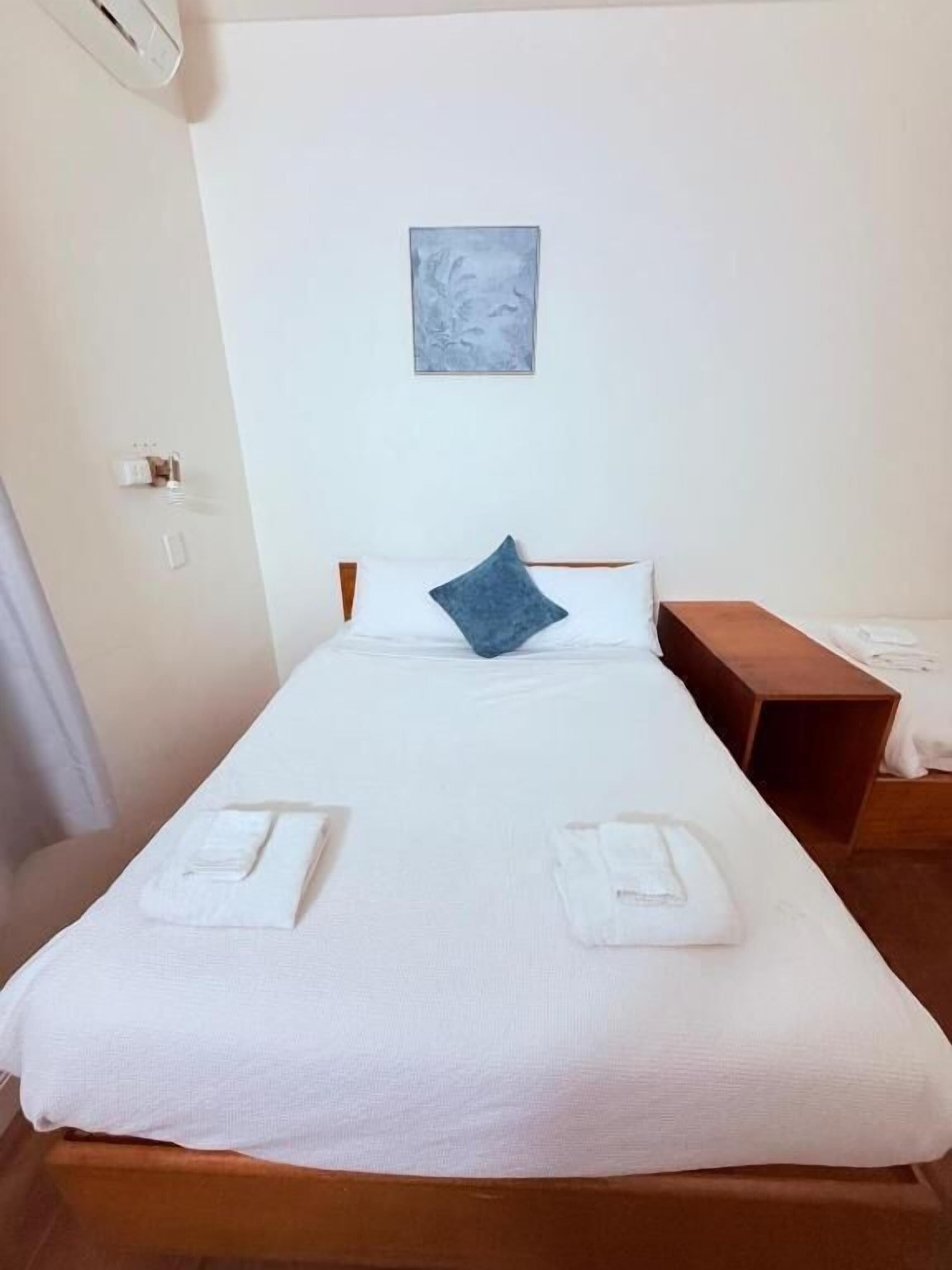Un lit simple avec du linge de lit blanc, un oreiller bleu et une tête de lit en bois. Il y a une petite table de chevet en bois sur la droite et un luminaire fixé au mur au-dessus du lit.