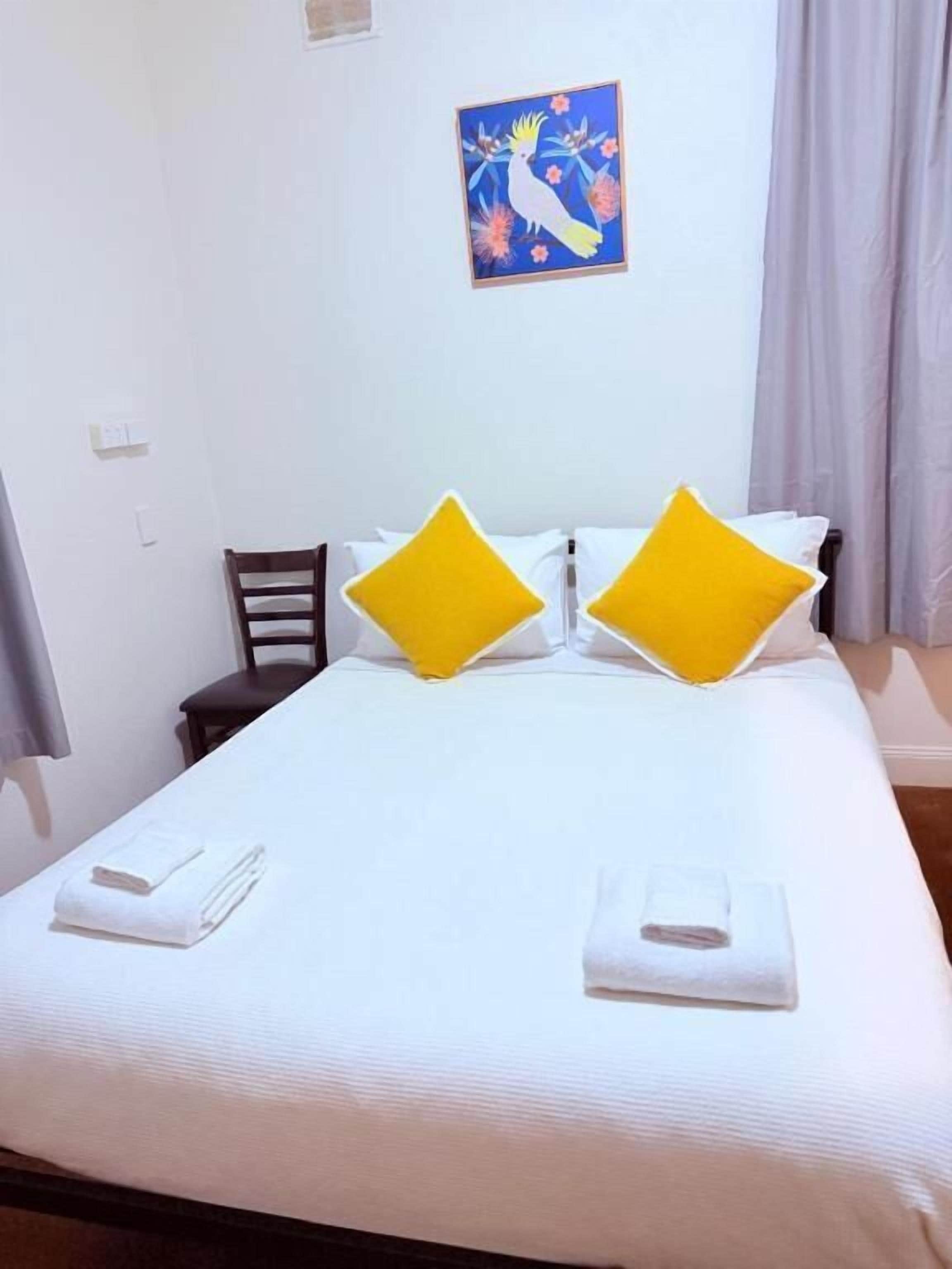 A neatly made bed with white bedding and two pillows, a chair, a painting on the wall, and a window with curtains.