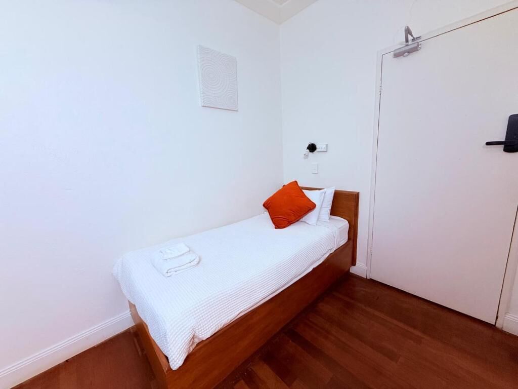 A single bed with a white mattress and orange pillow, a white towel, and a wooden headboard in a room with white walls and a wooden floor.