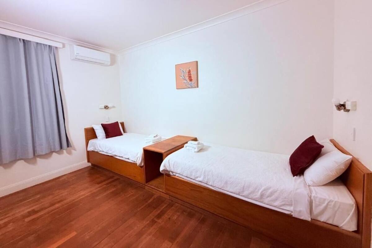 A room with two single beds, a wooden nightstand, a small table, a wall-mounted air conditioner, and a window with curtains.