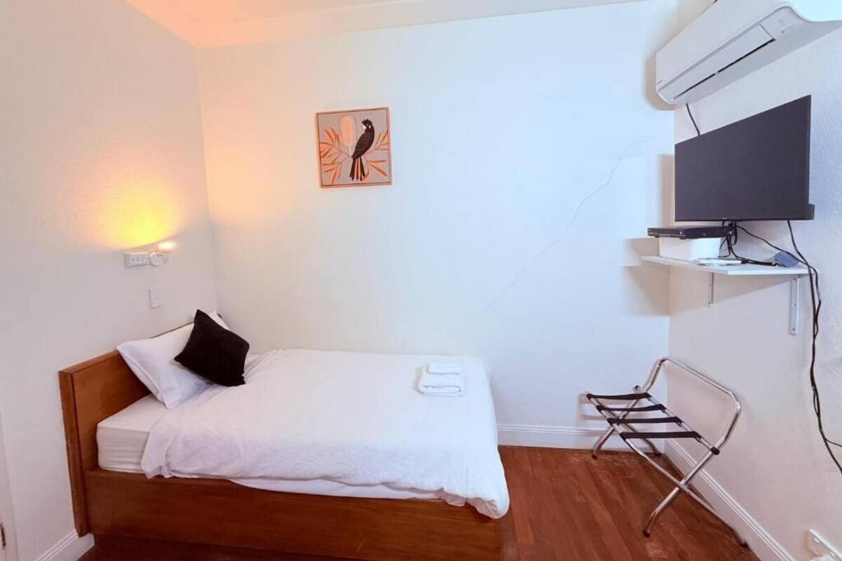A single bed with a wooden headboard, a wall-mounted TV, a small shelf with items, and a wall-mounted air conditioner.