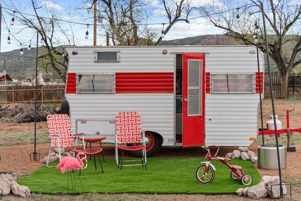 Una casa rodante con puerta roja, dos sillones de estampado rojo y blanco, una bicicleta roja y una pequeña estatua de flamenco rosa.