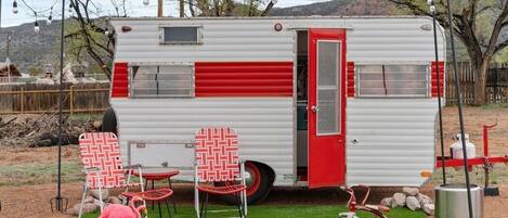 Una casa rodante con puerta roja, dos sillones de estampado rojo y blanco, una bicicleta roja y una pequeña estatua de flamenco rosa.