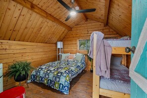 A wooden cabin with a sloped ceiling, a bunk bed, a single bed with a colorful quilt, a ceiling fan, and a small plant. - Bubble Gum Cottage | Vintage Charm | Near Cañon City Center (Canon City)