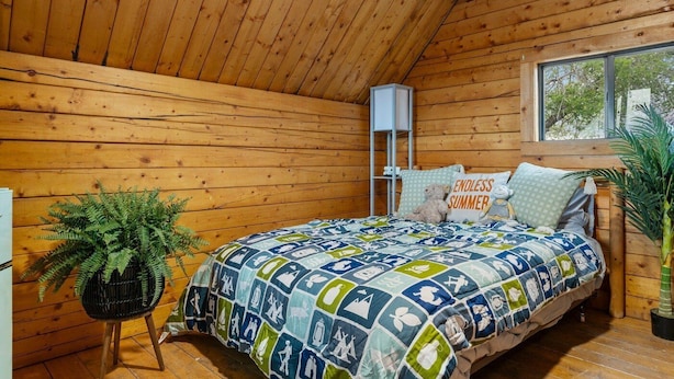 A cozy cabin room with a wooden ceiling, a bed with a colorful quilt, a small plant, and a window. - Cadillac Cottage Retreat | 1 Unit | Near Cañon City Center (Canon City)