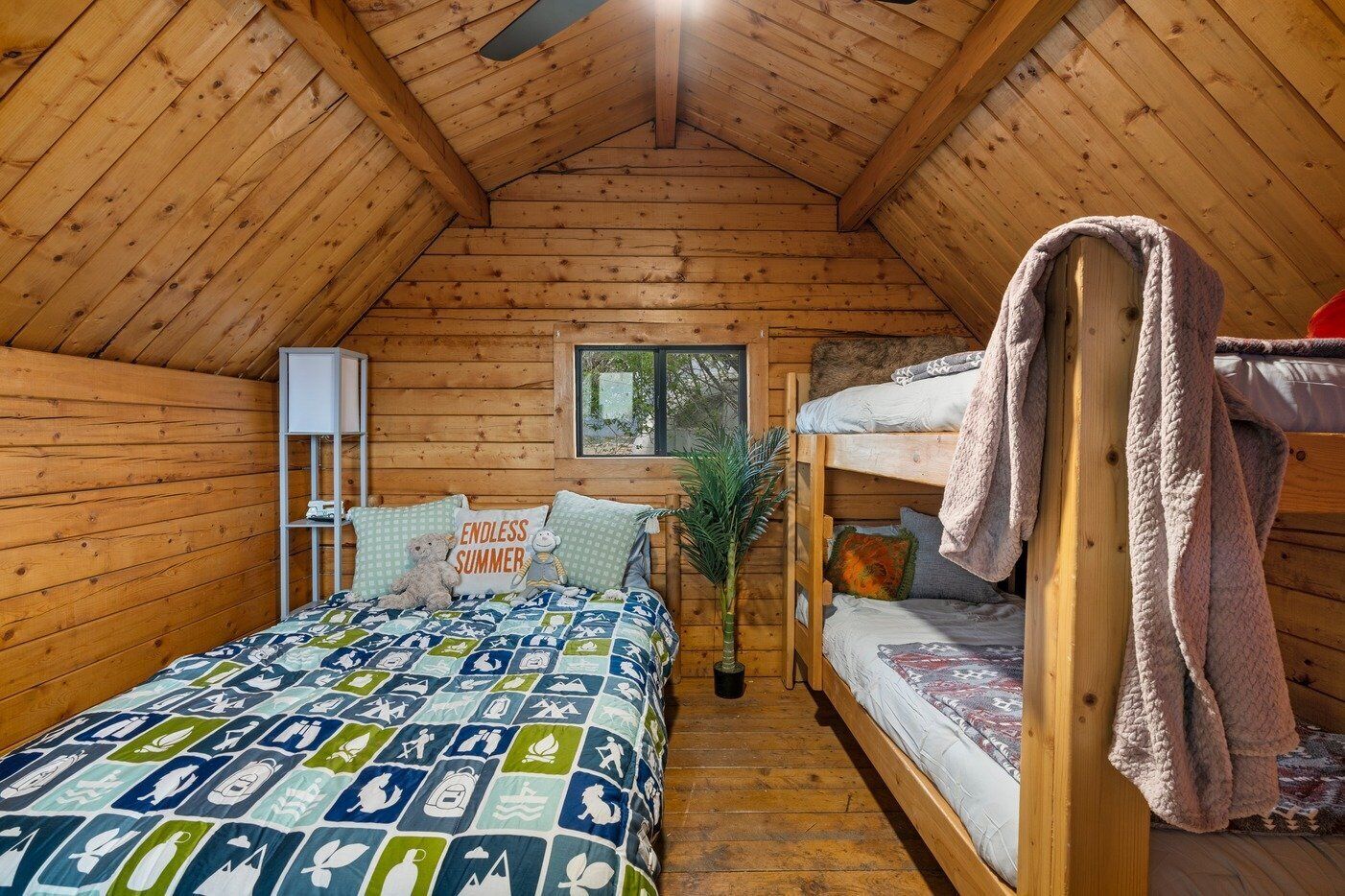 A cozy cabin with a wooden interior, a bunk bed, a bed with a patterned quilt, a small plant, and a window with a view.