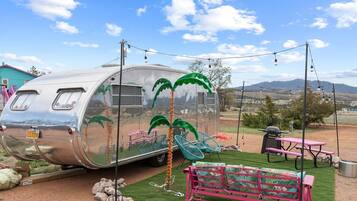 A vintage trailer with palm tree artwork, a picnic table, and a grill set up in a rural outdoor area.