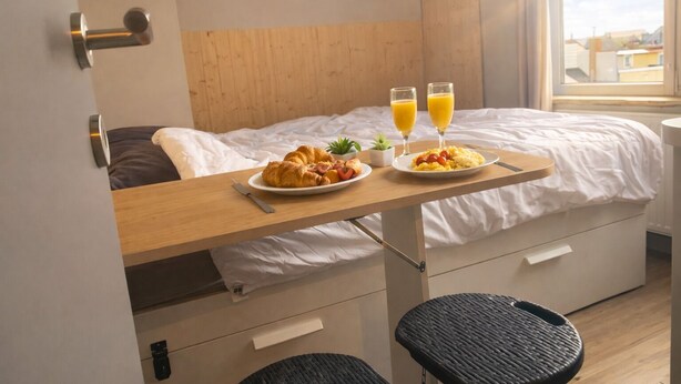 Una cama con cabecera de madera, una mesita de noche con desayuno, una ventana con vista a la ciudad y una pequeña planta en maceta.