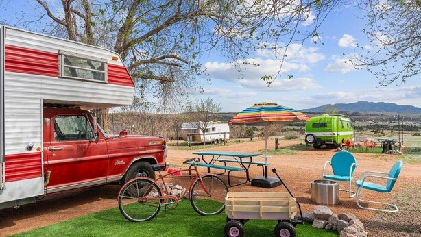 Una autocaravana con exterior rojo estacionada en un césped, una mesa de picnic, una bicicleta y un remolque verde al fondo.