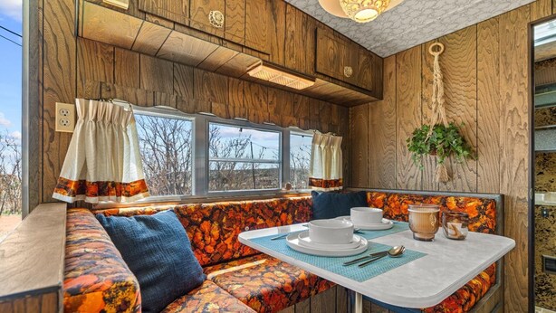 Ein gemütliches Interieur mit einer Holzwand, einem Fenster mit Vorhängen, einer Bank und einem Tisch, der mit Tellern und Tassen gedeckt ist.